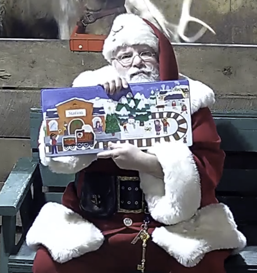 Santa sitting in green chair in front of reindeer holding book open facing the camera